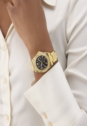 Goldfarbene Armbanduhr mit schwarzem Zifferblatt und Kristallakzenten, getragen am Handgelenk einer Person in einem weißen, langärmeligen, knöpfbaren Hemd.