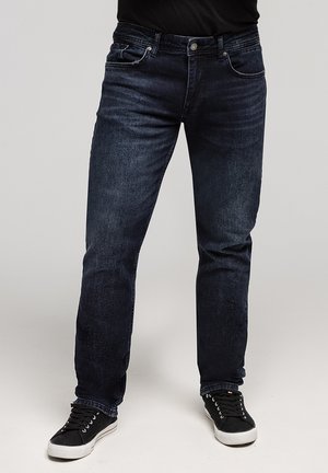 Osoba ubrana w ciemnoniebieskie dżinsy i czarne trampki z białymi podeszwami, stojąca na tle jasnoszarym.