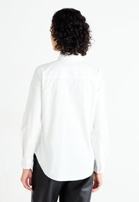 Camisa blanca de manga larga con cuello abotonado, corte entallado y dobladillo curvado, combinada con pantalones negros, que muestran una textura de tela suave.