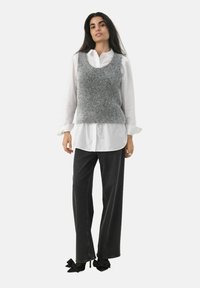 Grijze fuzzy gebreide vest over een witte blouse met kraag, gecombineerd met donkere wijde broek en zwarte hakken met strikaccenten.