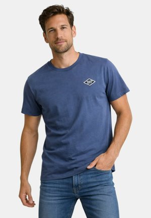 Homme portant un t-shirt bleu à manches courtes avec un petit logo blanc sur le côté gauche de la poitrine et un jean bleu, debout avec une main dans la poche.
