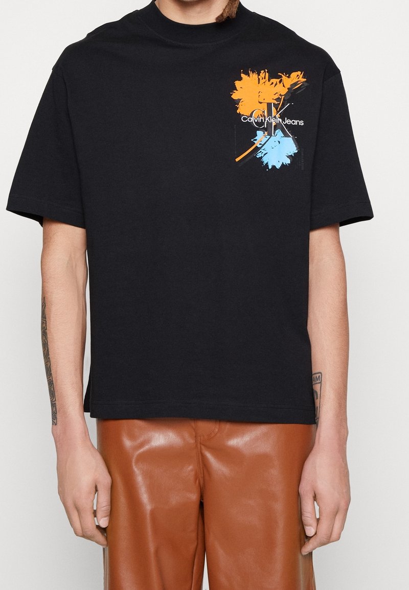 Person trägt ein schwarzes Calvin Klein Jeans T-Shirt mit einem orange-blauen Blumenmotiv auf der Brust, kombiniert mit braunen Lederhosen.
