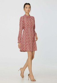 Femme portant une robe longue à manches longues rouge à fleurs et des sandales à talons beiges, se tenant devant un arrière-plan clair.