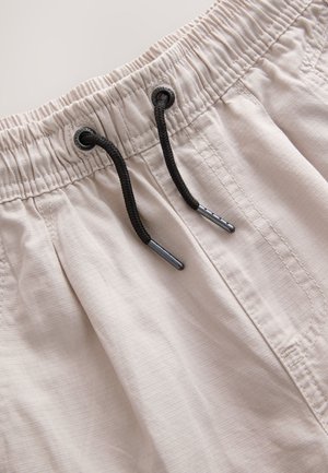 Shorts de bain beige avec une ceinture élastique et un cordon noir, présentant un tissu texturé avec de subtiles lignes verticales et des détails de couture.