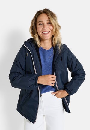Veste coupe-vent marine avec une capuche, des poignets élastiques et une fermeture éclair à l'avant. Associée à un pull bleu et un pantalon blanc, de coupe décontractée.