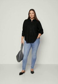 Camicia nera con bottoni, jeans skinny blu chiaro, ballerine nere e borsa grigia. Modella in piedi con una mano in tasca, sfondo neutro.