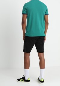 Tyrkysové polo tričko s námořnickými detaily, černé šortky a černé tenisky se zelenými akcenty. Bílé ponožky jsou viditelné nad kotníkem.