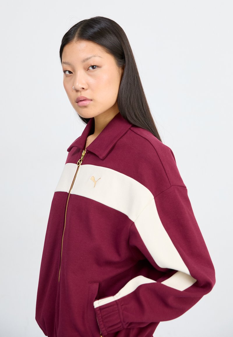 Burgunderfarbene Zip-Jacke mit cremefarbener Streifen, geripptem Kragen und Bündchen. Verfügt über ein goldenes Logo auf der linken Brust und seitliche Taschen. Weicher, strukturierten Stoff.