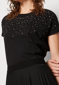 Černé triko s krátkým rukávem a kulatým výstřihem, zdobené rozptýlenými kovovými flitry a hladkým povrchem. Akcentováno žebrovaným pasem.