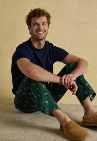 Tumši zilo kokvilnas T-kreklu un zaļos pidžamu bikses ar rotaļīgu sīku figūru rakstu. Brūni zamšādas klogs ar aizmugurējo siksniņu.