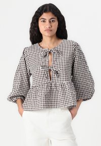 Blusa peplum a quadri marroni e bianchi con scollatura a oblò, fiocchi in raso al collo e maniche a palloncino. Tessuto in cotone liscio.