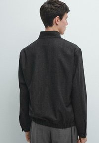 Czarna, teksturowana kurtka z kołnierzem, elastyczną talią i mankietami na guziki. Widok od tyłu pokazuje luźny krój i subtelny wzór jodełkowy.