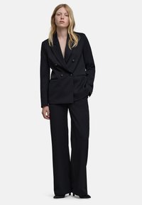 Vrouw in een zwarte dubbelbreasted blazer en wijde broek, staand met één hand in de zak tegen een effen achtergrond.