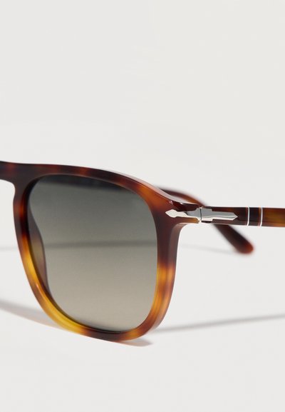 Καφέ γυαλιά ηλίου με καβούρι και σκούρα φακούς με διαβάθμιση, και ασημένιο λεπτομέρειες άρθρωσης σε σχήμα βέλους σε λευκό φόντο.