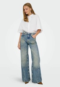 Witte oversized blouse met korte mouwen, gecombineerd met ruime lichtblauwe denim jeans. De jeans hebben een vervaagde wasbeurt en een stevige tailleband.