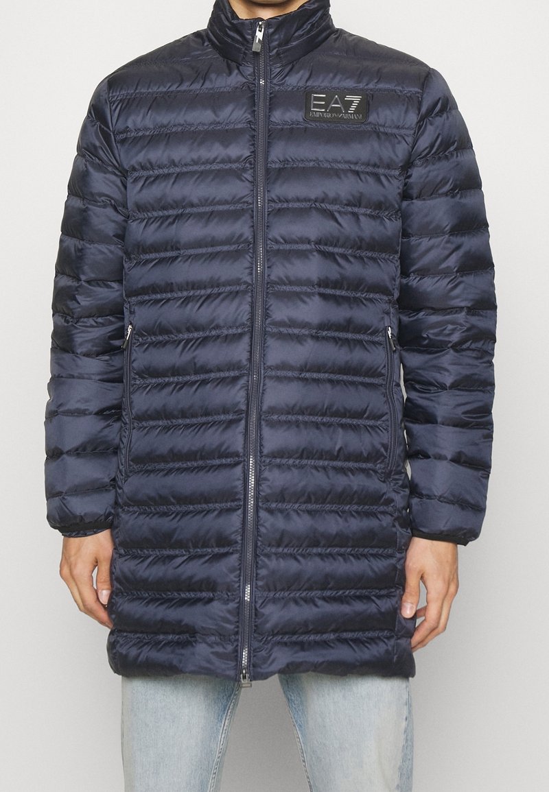 Navy pufferjacka med horisontellt quiltat mönster, hög krage, frontdragkedja och EA7-logopatch på övre vänstra sidan.