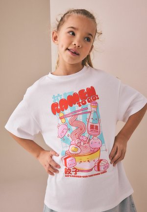 Ung pige iført en hvid T-shirt med farverigt tegneserie-design af en ramen-skål, en flaske soja og spisepinde, smilende og poserende med hænderne på hofterne.