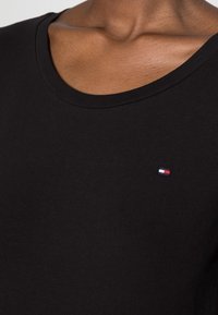 Černé bavlněné tričko s kulatým výstřihem; na levém prsu je malé vyšívané logo v červené, bílé a modré barvě.