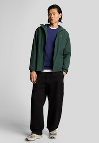 Giacca leggera verde con cappuccio, felpa blu, pantaloni larghi neri e sneakers con motivo. Logo distintivo sul petto.
