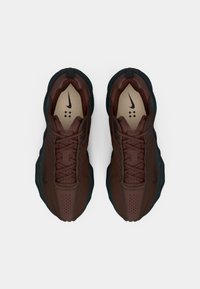 Tênis Nike com um cabedal castanho escuro, detalhes texturizados, sola preta e pormenores perfurados, com um logo na língua plana.