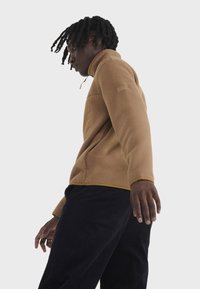 Brauner Fleece-Pullover mit hohem Kragen, Reißverschluss und Seitentaschen. Hat eine strukturierte Oberfläche und einen kontrastierenden gelben Besatz am Kragen.