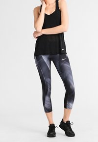Czarny sportowy top na ramiączkach z dekoltem w serek i luźnym krojem, zestawiony z czarnymi legginsami w szaro-czarny abstrakcyjny wzór. Całość uzupełniają czarne buty sportowe.
