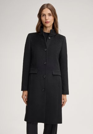 Manteau en laine noire avec un col montant, fermeture à boutons et poches latérales. Présente une finition texturée et une coupe ajustée, s'étendant jusqu'à mi-cuisse.