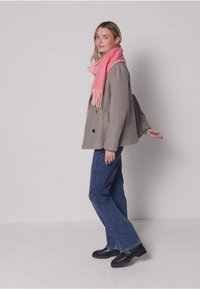 Veste grise en mélange de laine, écharpe rose à franges, jean bleu et mocassins noirs. Le mannequin pose de profil sur un fond clair.