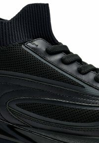 Primer plano de un zapato deportivo negro con collar de punto en el tobillo, paneles de malla y cuero, y cordones redondos atados con fuerza.