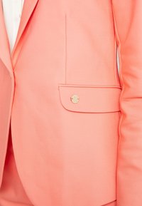 Blazer rose corail avec poche à rabat ornée d'un bouton rond en or et un revers visible sur le côté gauche.