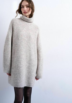 Femme portant une robe-pull col roulé ample gris clair à manches longues et des collants noirs, sur fond blanc uni.