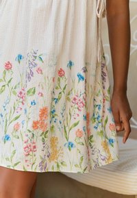 Lahka obleka z teksturo v kremni barvi z cvetličnim vzorcem, ki vključuje rožnate, modre in oranžne cvetove ob robu. Prilagodljive vrvi.