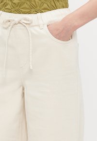 Light beige, relaxed-fit trousers with an elastic waistband, drawstring detail, and side pockets; fabric appears soft and slightly textured.