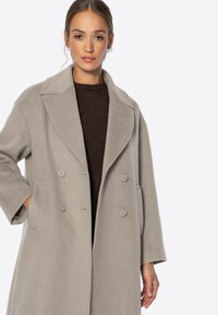 Doppelreihiger, hellgrauer Mantel aus weichem Stoff, mit großen Revers, zwei Fronttaschen und Knopfakzenten.