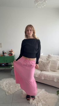 Jeune femme portant un haut noir à manches longues et une jupe rose, debout dans un salon lumineux avec un canapé blanc et une armoire verte.
