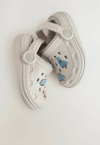 Sabots beiges avec une surface texturée, ornés de charms en forme de dinosaure bleus et roses, et de double sangles latérales pour un maintien sécurisé.