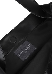 Interno di una borsa a tracolla in pelle nera, con rivestimento in tessuto, un'etichetta prominente con la scritta "Tucano Milano 1985" e un dettaglio di chiusura a scatto.