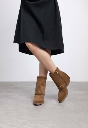 Botas de tobillo de ante marrón con un acabado texturizado, que cuentan con una cremallera lateral y tachuelas decorativas cerca del tobillo. La altura del tacón es moderada.