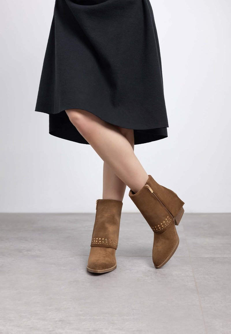 Botas de tobillo de ante marrón con un acabado texturizado, que cuentan con una cremallera lateral y tachuelas decorativas cerca del tobillo. La altura del tacón es moderada.