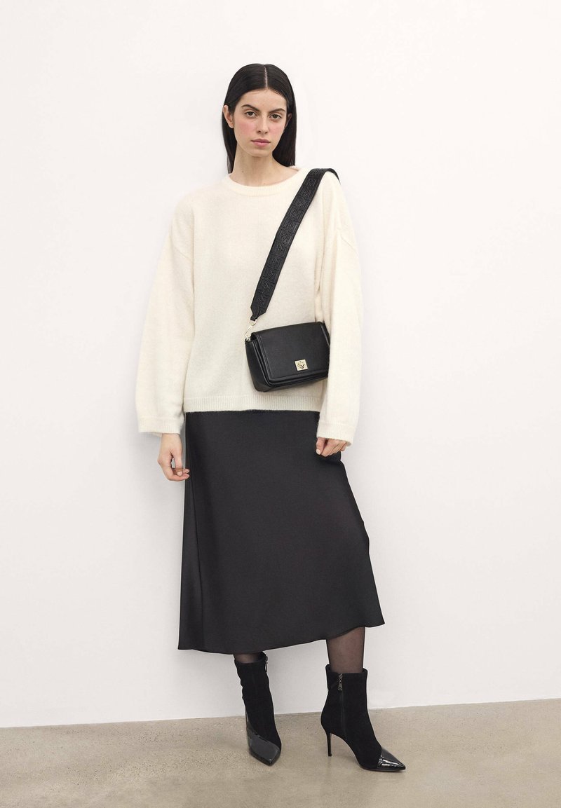 Maglione crema oversize, gonna midi nera in satin, stivali neri alla caviglia e borsa a tracolla nera con dettagli in rilievo sulla tracolla.