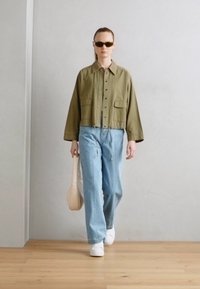 Veste verte courte avec deux poches avant, jeans larges bleu clair, baskets blanches et sac fourre-tout beige. Le modèle porte des lunettes de soleil noires.