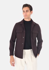 Giacca di lana marrone con chiusura a bottoni, due tasche frontali e colletto a punta. Indossata sopra una camicia nera e pantaloni bianchi.