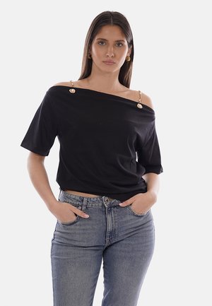 Donna che indossa un top nero scollato con spalline a catena dorata e jeans blu, in piedi con le mani nelle tasche davanti a uno sfondo neutro.