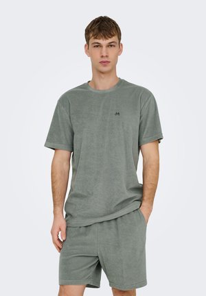 Jonge man draagt een simpel grijs badstoffen t-shirt en bijpassende short, staat met één hand in de zak tegen een effen achtergrond.