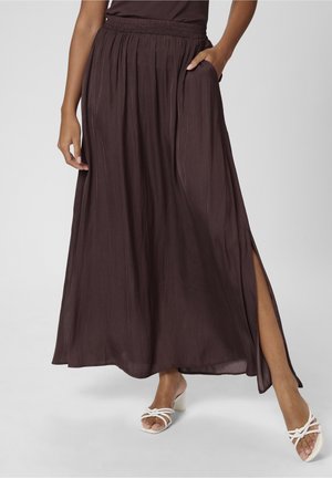 Kobieta w długiej, ciemnobrązowej spódnicy maxi z bocznym rozcięciem i kieszeniami, w białych sandałach na obcasie, stojąca na jednolitym tle.