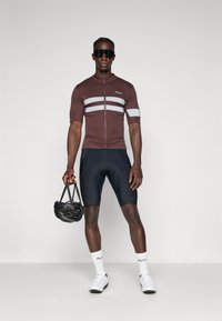 Cyklistický dres v tmavě hnědé barvě se stříbrnými pruhy, krátké rukávy a přední zip. Černé kraťasy, bílé ponožky a bílé cyklistické boty, držící černou helmu.