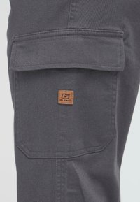 Dark gray fabric cargo pocket with flap and small brown leather patch labeled "Blend" on the side of pants.