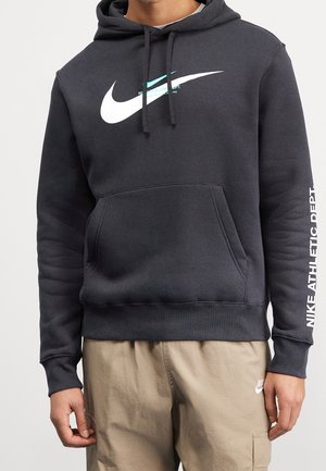 Mężczyzna noszący czarną bluzę z kapturem Nike z białym logo swoosh i napisem na piersi oraz rękawie, w połączeniu z beżowymi szortami Nike.