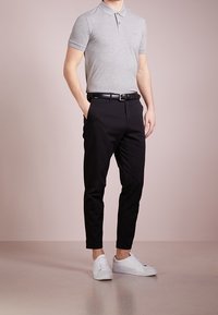Polo shirt gris à manches courtes avec une coupe ajustée, associé à un pantalon noir fuselé et des baskets blanches, se tenant sur un sol neutre.