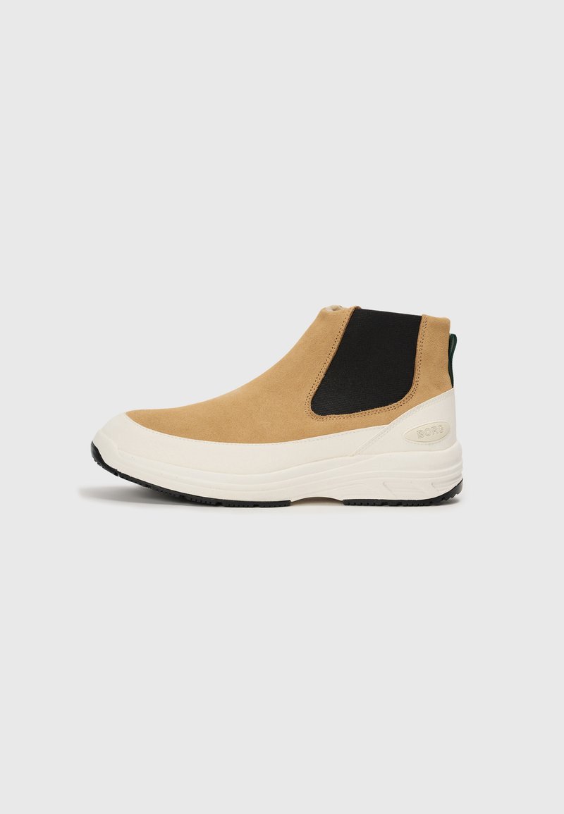 Botín de ante color marrón con paneles elásticos negros, suela de goma blanca y un acento verde en el talón. Diseño elegante con líneas limpias.